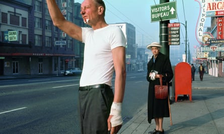 Man With bandage, 1968, by Fred Herzog. The man seems to be hailing an approaching bus, while an elderly woman looks on quizzically, keeping her distance. The image is almost Hitchcockian.