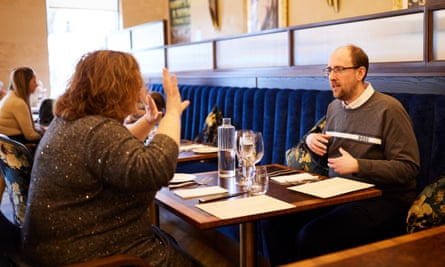 A young, balding man with glasses, wearing a grey top over a white shirt, and an older woman with curly red hair and glasses, wearing a grey sparkly top, sitting at a wooden restaurant table talking