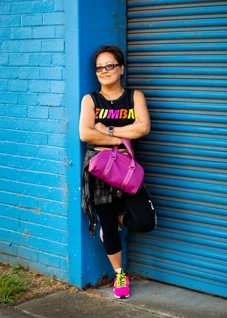 A middle-aged Asian woman in a black singlet with the word “Zumba” in neon letters, stands with her arms crossed against a blue wall.