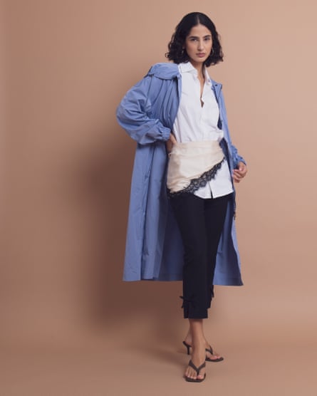 A woman poses in a studio wearing a light blue coat over a white shirt, waist scarf, black capri pants and heeled sandals