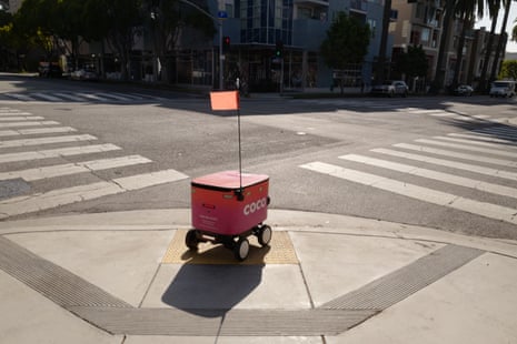 A pink Coco delivery robot with an orange flag waits at a street corner by a crosswalk