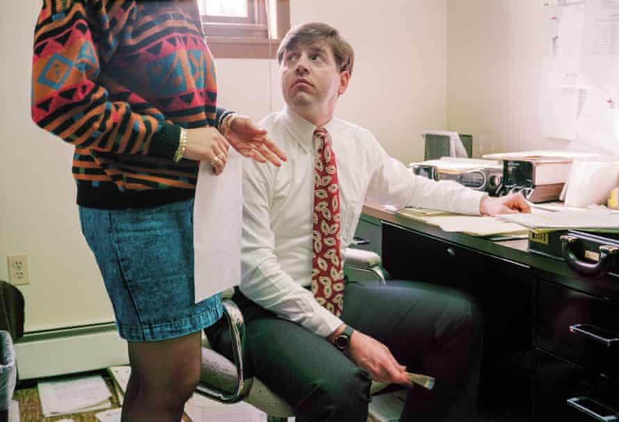 Man sitting at desk in office, looking at woman in denim skirt and patterned sweater