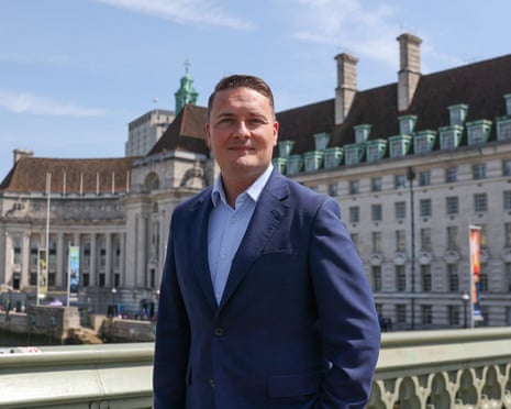 Wes Streeting in a navy blazer and blue shirt stands on Westminster Bridge with historic buildings behind him