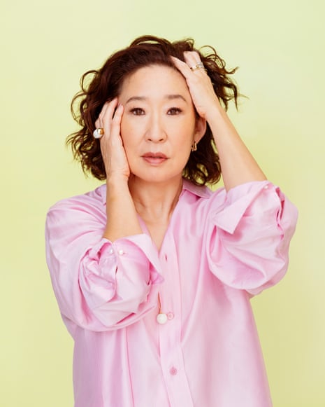 Sandra Oh in a pink shirt holds both hands to her head against a pale yellow background