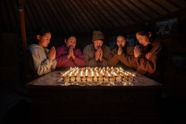 People clasp their hands in prayer around a table covered in dozen of small oil lamps