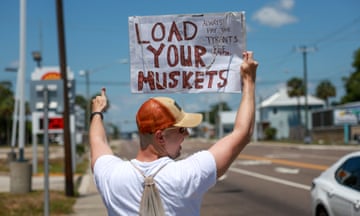Young white man wearing American flag hat holds up white sign beside busy street; back of sign says Load Your Muskets.