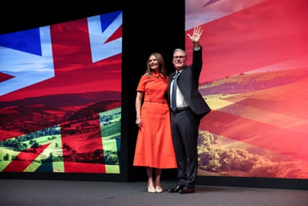 Keir and Victoria Starmer stand on a stage in front of a projection including the union jack flag; she wears a red dress and he is waving
