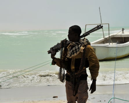 An armed man stands on a beach with a boat in the background