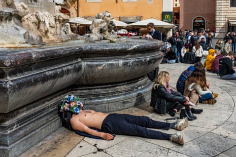 he is bare-chested with black jeans and dirty white trainers, and has a flower-patterned shirt over his face. He rests his head on a black rucksack.