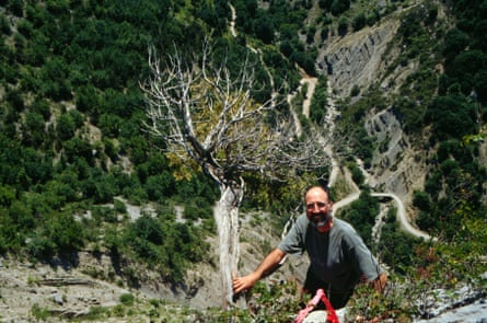 Vue d'en haut de Doug Larson sur une falaise tenant un arbre avec une longue chute derrière lui avec une route en lacets qui serpente autour de la montagne.