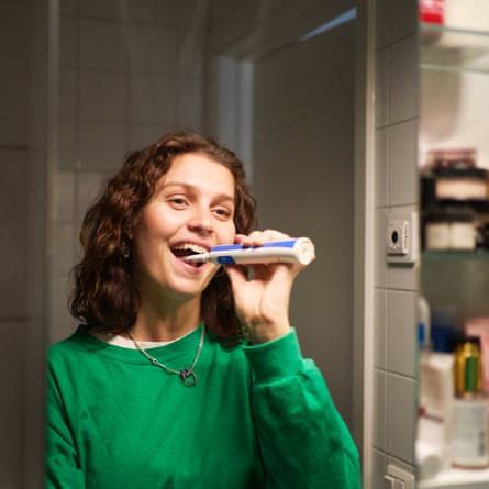 Smiling young woman brushing teeth with electronic toothbrush while looking in mirror at bathroom
