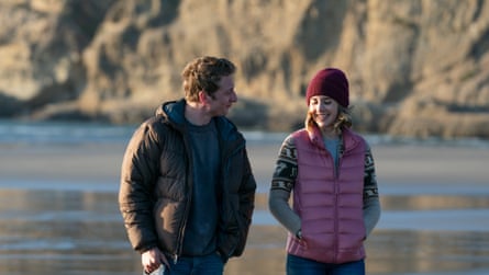 They walk on a beach, her in a pink gilet, him in a puffer jacket
