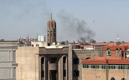 Black smoke rises into a greyish-blue sky above buildings. The cityscape includes one structure with an elaborately carved stone facade, tiled roofs, and the tall minaret of a mosque.