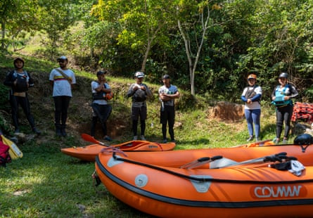 A small group of people wearing jeans and T-shirts stand in a loose circle around a bright orange river raft and a hard-sided orange kayak.