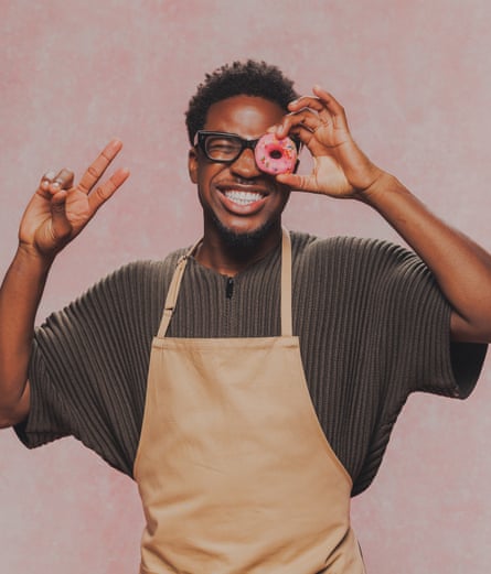 A young black man holds up a peace sign with one hand while holding a pink donut over one bespectacled eye with the other.