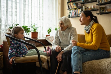 An elderly white-haired woman sits in a living room between a toddler girl and a young woman who looks like the girl’s mother.
