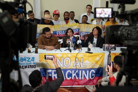 Two women and a man sit at a table in front of cameras speaking into microphones, while people behind them hold signs which read ‘No toFracking’in Spanish,.asdoes a banner covering the front of the table.