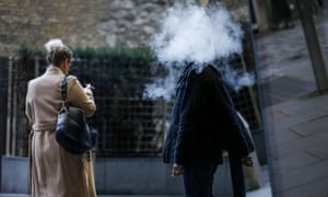 A pedestrian stands surrounded in a cloud of vapour after exhaling from a vape device as a pedestrian smoking a cigarette passes in London