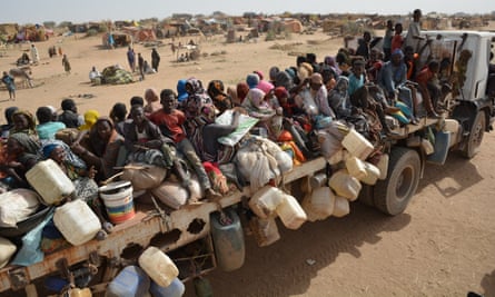 A large number of people sit on the back of a crowded truck with their belongings, in an arid landscape