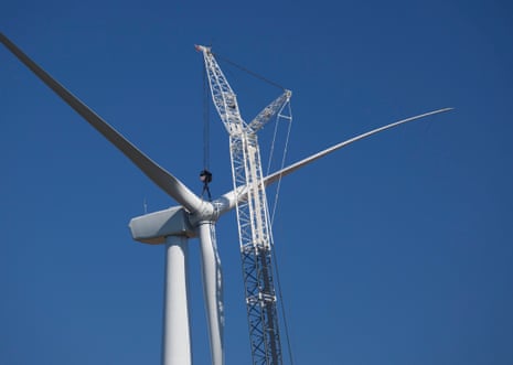 A crane lifts a wind turbine blade beside an installed turbine during wind farm construction