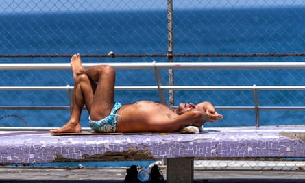 A man in swimming trunks lies along the Beirut promenade in front of the sea, stretched out, legs crossed, arms folded beneath his head