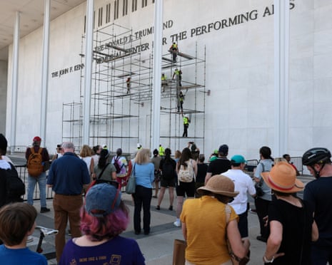 A crowd looks on as lettering referencing Donald Trump is removed from the facade of the John F. Kennedy Center for the Performing Arts.