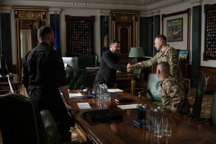 Mykhailo Drapatyi shakes hands with Volodymyr Zelenskyy in a formal meeting room. They stand by a long varnished wood table and chairs covered in green leather. Zelenskyy wears black and Drapatyi is in military camouflage. Two other men are present with their backs to the camera.