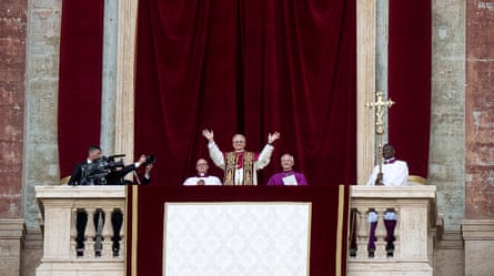 Pope Leo XIV waving from the balcony of St Peter’s Basilica with two men standing either side of him.
