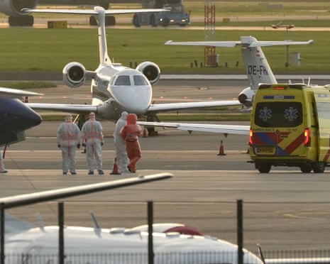 People in hazmat suits stand near a small aircraft on a tarmac next to a yellow ambulance.
