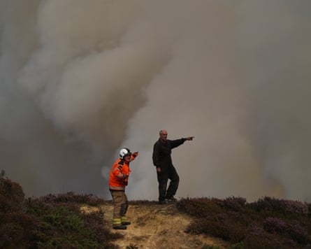A firefighter in hi-vis and another man stand on the moor and point towards an area of billowing smoke that obscures the view beyond the moor