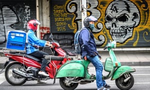 A man riding a scooter waits on a traffic light in front of graffiti on a store’s shutters in downtown Bangkok.