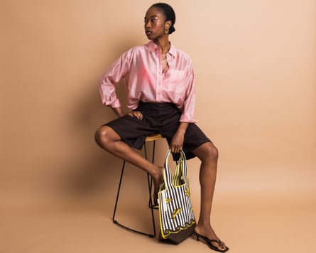 A model in a pink shirt, black shorts and black shoes with a black-and-white striped bag, sitting on a stool.