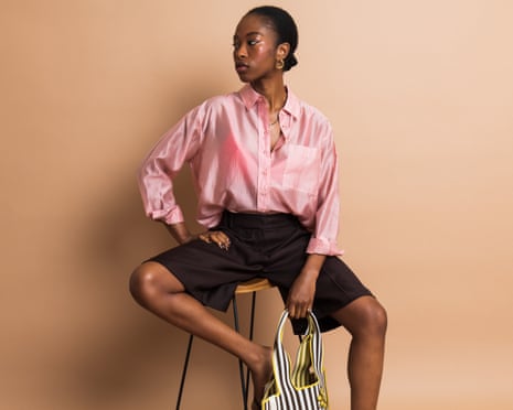 A model in a pink shirt, black shorts and black shoes with a black-and-white striped bag, sitting on a stool.
