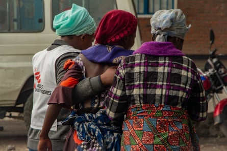 Two women, one with an MSF bib support a third woman as they walk to a building