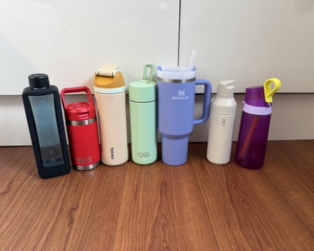 Seven water bottles of all different colours in a line on a wooden worktop in front of white cupboards