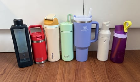 Seven water bottles of all different colours in a line on a wooden worktop in front of white cupboards