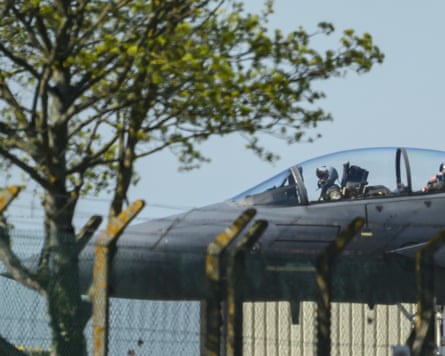 Nose of plane with pilot visible in cockpit on ground behind fence/tree