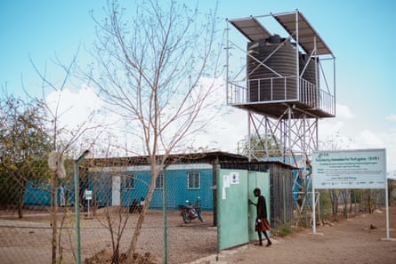 A person walks through a gate outside a single level building with a large water tank suspended on scaffolding.