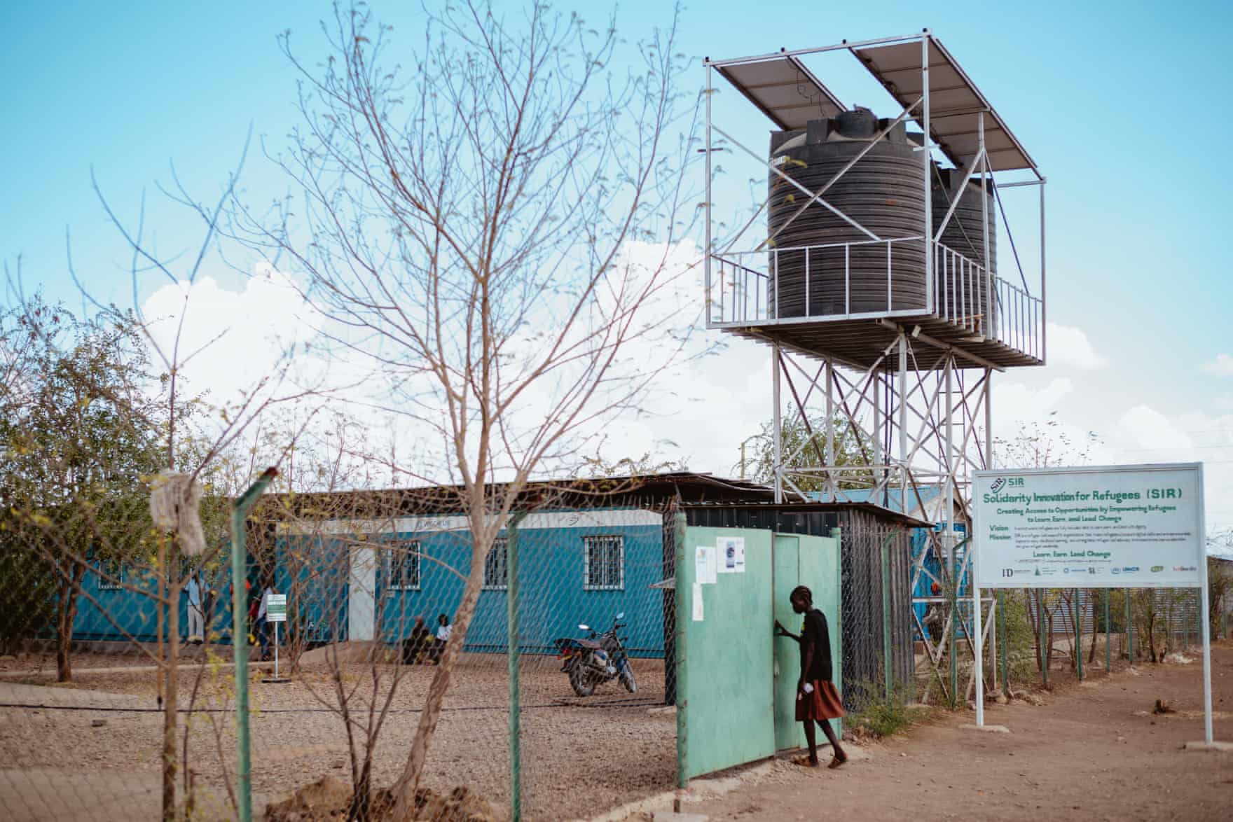 A person walks through a gate outside a single level building with a large water tank suspended on scaffolding.