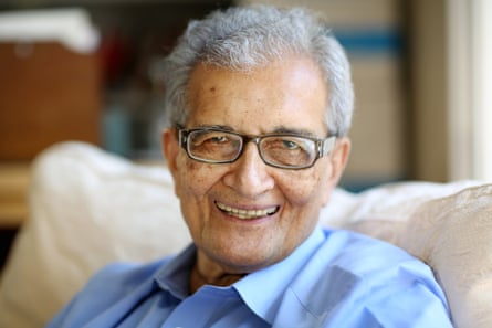A man sitting on a cream sofa wearing a blue shirt and glasses smiles at the camera