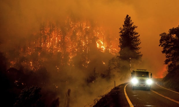 A firetruck drives down a road with its lights on as a mountain in the background can be seen with trees ablaze from a forest fire.