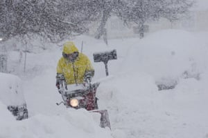 A person in a yellow coat with hood up uses a blower to clear snow