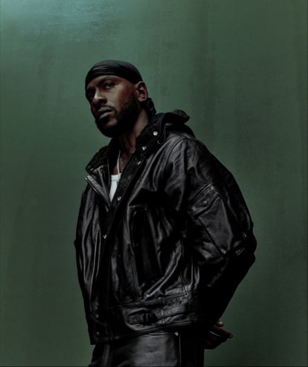 Skepta in a black leather jacket and skullcap looks away from the camera pensively