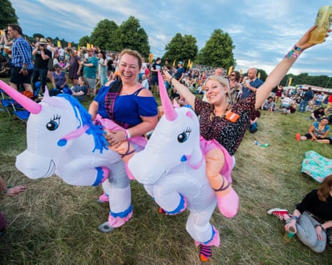 Two women in inflatable unicorn costumes smile and pose at an outdoor festival with crowds in the background