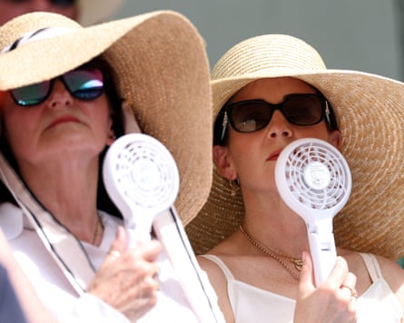Two women wearing wide-brimmed sun hats use handheld fans to blow air on their faces
