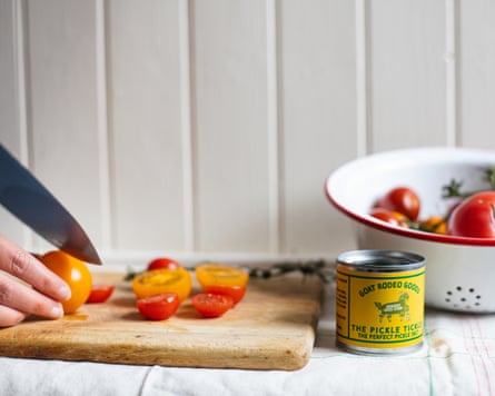 A person chopping tomatoes on a wooden chopping board with a tin of Goat Rodeo Goods in view.