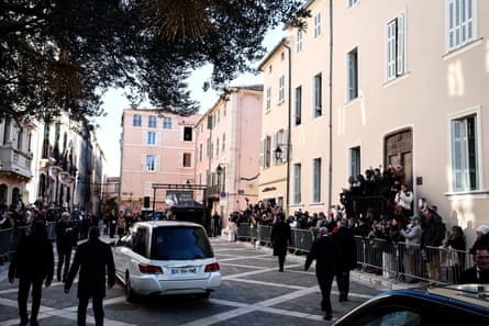 A white Mercedes hearse travels down a stone-paved street as people including photographers look on from behind metal barriers. Men in dark jackets walk behind and around the hearse. The street is lined with tall, pale pink and peach-coloured buildings with white shutters, typical of the south of France.