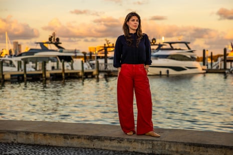 A woman wearing a black shirt and red pants stands for a portrait by a yacht marina