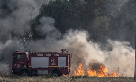 Israeli firefighters work to extinguish a fire: a red fire engine is seen next to billowing flames in a field; clouds of smoke rise high behind it. 