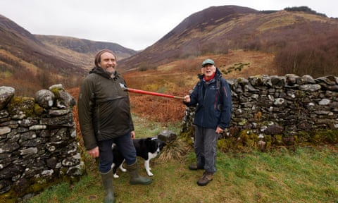 Two men dressed in outdoor gear stand with a dog by an old stone wall in the Carrifran valley.